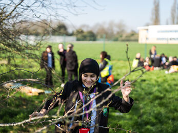 A schoolgirl reaching for the branches of a tree, smiling. Behind her can be seen a group of her classmates, sat on lush green grass