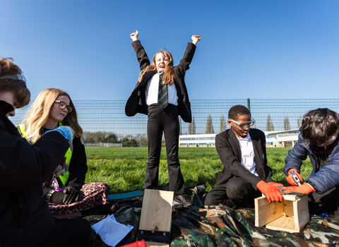 A group of secondary school students sat on the grass outside making bird boxes. One young person is stood up, holding their arms triumphantly up in the air